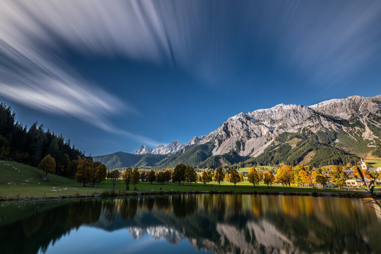 Spiegelung verschneiter Berge und herbstlicher Bäume in einem ruhigen See unter blauem Himmel. | © Oliver Richter Leica