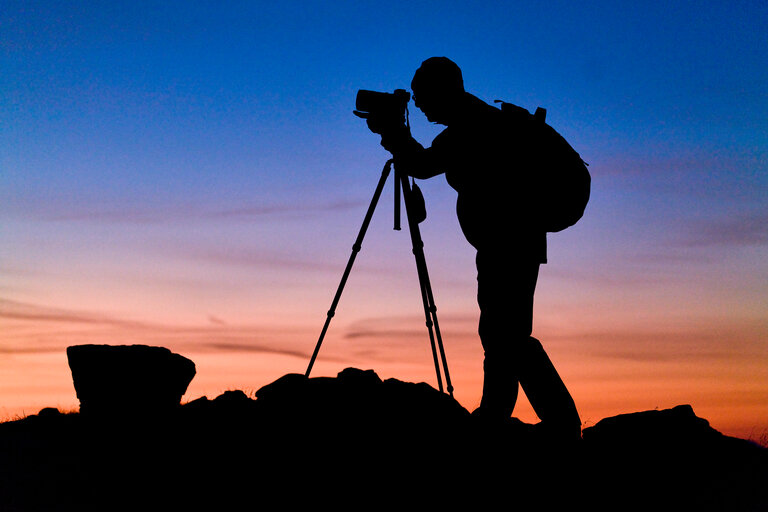 Das Foto zeigt die Silhouette einer Person mit Kamera und Stativ, die bei Sonnenaufgang oder Sonnenuntergang in der Natur fotografiert. | © Oliver Richter Leica